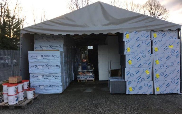 Pannelli X-FOAM e lastre isolanti imballate sotto un gazebo grigio nel deposito Tecnoedil, con secchi bianchi su pallet