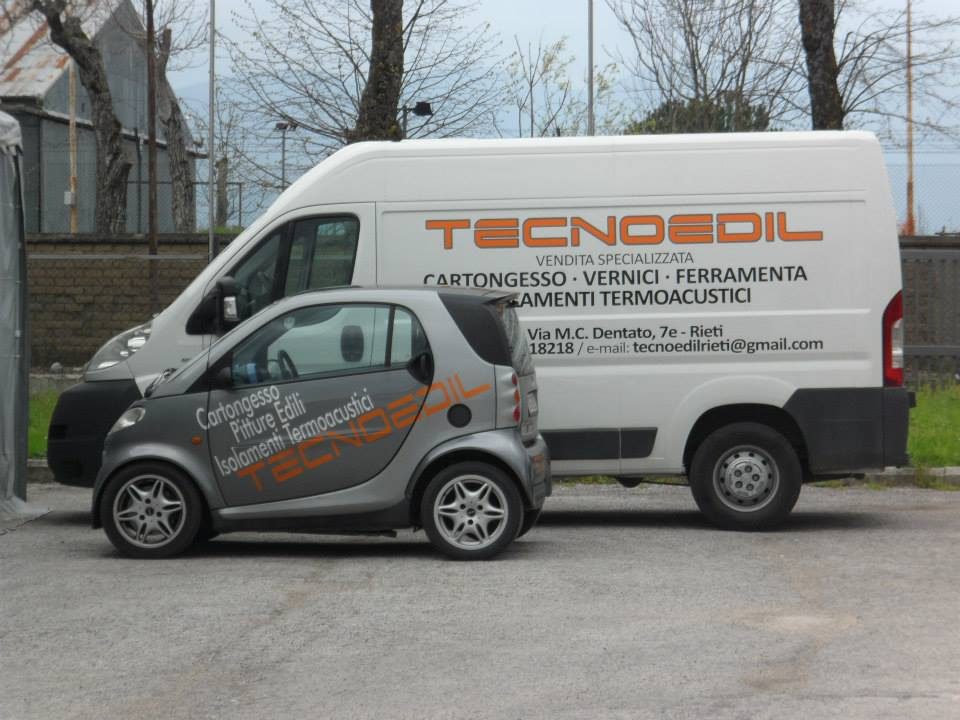 Furgone bianco e citycar grigia con scritte Tecnoedil arancioni, «Cartongesso · Vernici · Ferramenta», in un piazzale